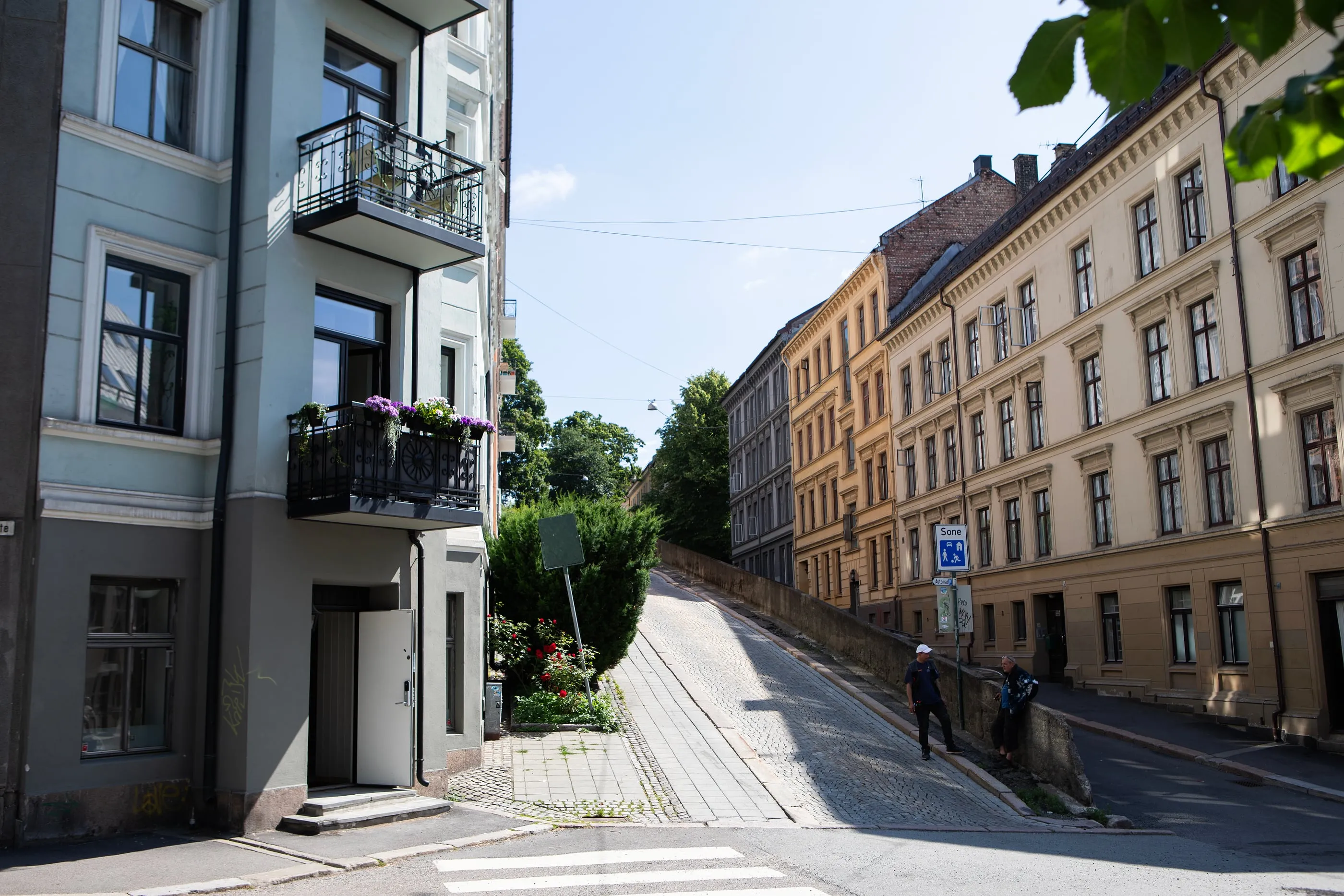 Oslolainen kulmatalo, jonka julkisivussa on kaksi mustaa teräsparveketta kukkineen, mukulakivikadun varrella